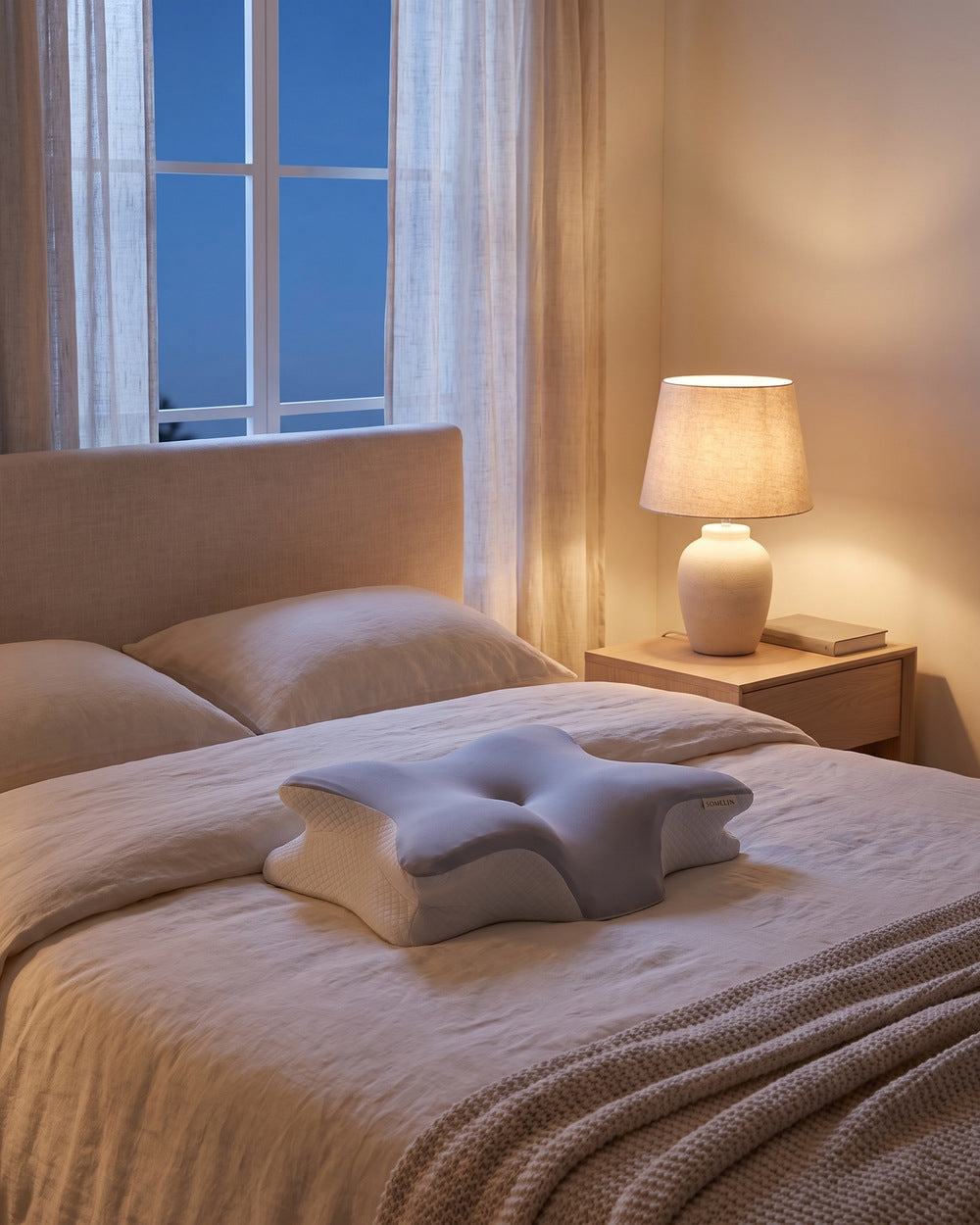 Chambre au crépuscule, l'oreiller Somélya sur un lit en lin crème, lampe de chevet allumée